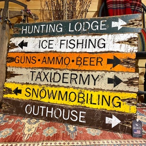 Puede incluir: Un letrero r&uacute;stico de madera con m&uacute;ltiples tablones horizontales, cada uno pintado de un color diferente y con texto blanco y flechas negras. El letrero incluye "HUNTING LODGE", "ICE FISHING", "GUNS &bull; AMMO &bull; BEER", "TAXIDERMY", "SNOWMOBILING" y "OUTHOUSE".