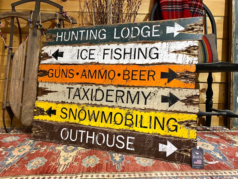 Puede incluir: Un letrero r&uacute;stico de madera con m&uacute;ltiples tablones horizontales, cada uno pintado de un color diferente y con texto blanco y flechas negras. El letrero incluye "HUNTING LODGE", "ICE FISHING", "GUNS &bull; AMMO &bull; BEER", "TAXIDERMY", "SNOWMOBILING" y "OUTHOUSE".