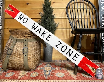 May include: A long, rectangular wooden sign with a white background and black text that reads NO WAKE ZONE. The sign features bright red painted ends with distressed, weathered wood grain visible at the tips. A small tag is attached to the bottom.