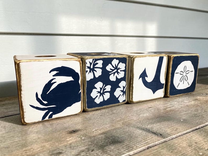 Four coastal tissue boxes with a distressed finish, each featuring a nautical or tropical design in navy blue and white. They display a crab, a hibiscus flower pattern, a ship anchor, and a sand dollar for beach and nautical decor.