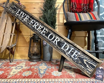 May include: A rustic wooden sign with a black background and white lettering that reads "Live Free or Die". The sign is framed in a dark wood frame and has a distressed look. The sign is leaning against a wooden chair and a wooden sled.