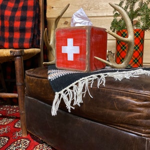 Rustic First Aid / Swiss Cross wood tissue box cover displayed in a ski lodge or cabin bathroom, perfect for beach houses, poolside guest rooms, cozy cabins, and lodge-style décor. Handcrafted from wood with natural texture, this vintage-inspired piece adds functional charm and nostalgic ski and lifeguard vibes