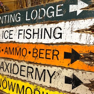 Puede incluir: Una pila de letreros de madera r&uacute;sticos con letras blancas y flechas negras. Los letreros indican direcciones a un refugio de caza, pesca en hielo, armas, municiones, cerveza, taxidermia y motos de nieve. Los letreros est&aacute;n pintados en varios colores.