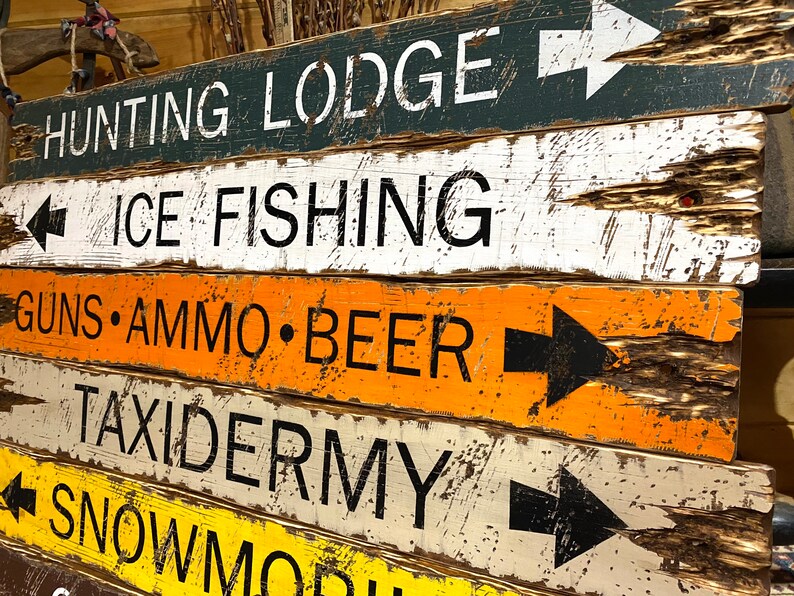Puede incluir: Una pila de letreros de madera r&uacute;sticos con letras blancas y flechas negras. Los letreros indican direcciones a un refugio de caza, pesca en hielo, armas, municiones, cerveza, taxidermia y motos de nieve. Los letreros est&aacute;n pintados en varios colores.