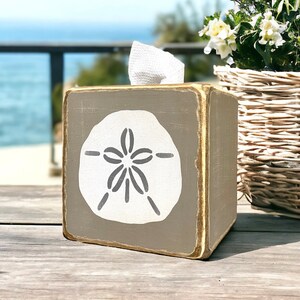 May include: A gray wooden tissue box cover with a white sand dollar design. The tissue box cover is sitting on a wooden table.