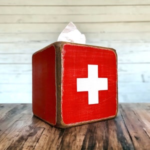 Rustic First Aid wood tissue box cover photographed in a studio setting, ideal for ski cabin décor, lodge bathrooms, beach houses, poolside spaces, and guest rooms. Handcrafted from wood with natural texture, showcasing vintage first aid design and rustic charm for functional home décor