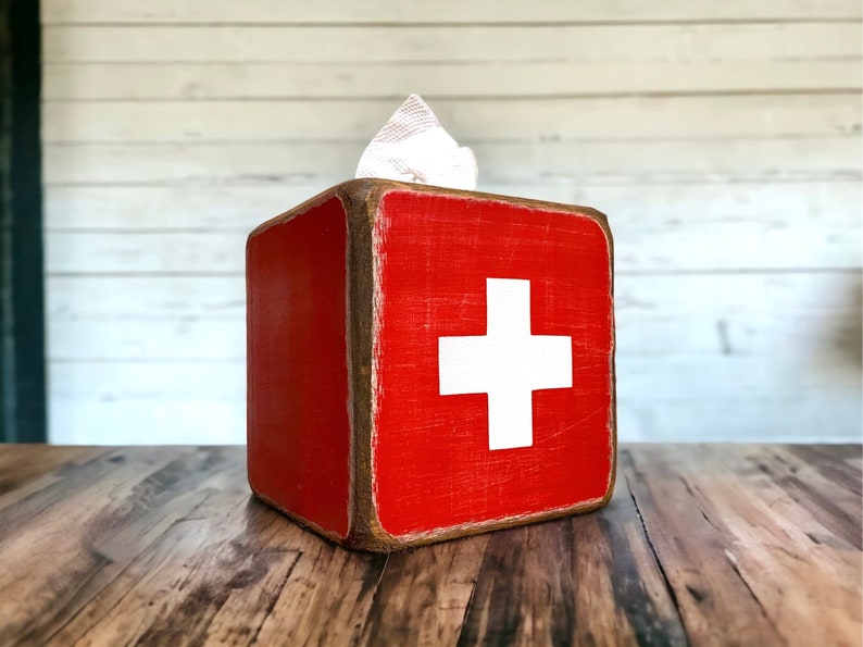Rustic First Aid wood tissue box cover photographed in a studio setting, ideal for ski cabin décor, lodge bathrooms, beach houses, poolside spaces, and guest rooms. Handcrafted from wood with natural texture, showcasing vintage first aid design and rustic charm for functional home décor