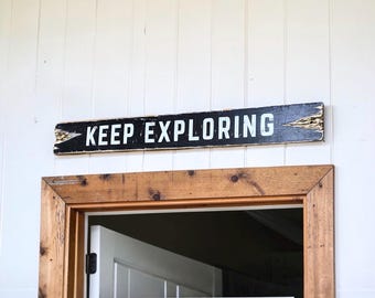 May include: A rectangular wooden sign with a distressed black finish and white lettering that reads KEEP EXPLORING. The sign features pointed ends and is mounted horizontally above a wooden door frame, displaying visible wood grain and wear.