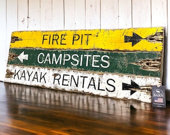Könnte beinhalten: Ein rustikales Holzschild mit drei Textzeilen. Die obere Zeile lautet "FIRE PIT" mit gelbem Hintergrund und einem schwarzen Pfeil, der nach rechts zeigt. Die mittlere Zeile lautet "CAMPSITES" mit grünem Hintergrund und einem schwarzen Pfeil, der nach links zeigt. Die untere Zeile lautet "KAYAK RENTALS" mit weißem Hintergrund und einem schwarzen Pfeil, der nach rechts zeigt. Das Schild ist verwittert und hat einen abgenutzten Look.