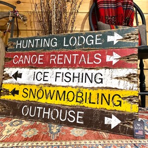 May include: Rustic wooden sign with five painted planks, each with a different activity and arrow pointing to the right. The activities are "Hunting Lodge", "Canoe Rentals", "Ice Fishing", "Snowmobiling", and "Outhouse".