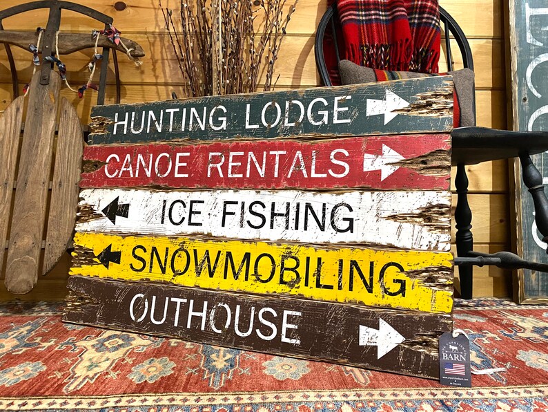 May include: Rustic wooden sign with five painted planks, each with a different activity and arrow pointing to the right. The activities are "Hunting Lodge", "Canoe Rentals", "Ice Fishing", "Snowmobiling", and "Outhouse".