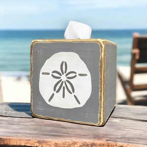 Gray sand dollar tissue box cover styled on a sandy beach with weathered coastal tones, ocean-inspired beach house decor ideal for seaside cottages, coastal bathrooms, vacation rentals, and relaxed salt-and-sand interiors, rustic distressed wood design