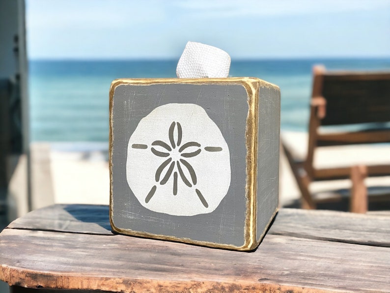 Gray sand dollar tissue box cover styled on a sandy beach with weathered coastal tones, ocean-inspired beach house decor ideal for seaside cottages, coastal bathrooms, vacation rentals, and relaxed salt-and-sand interiors, rustic distressed wood design