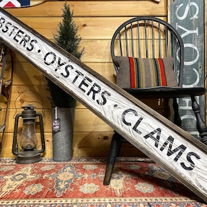 May include: A rustic wooden sign with a black frame reads "Lobsters, Oysters, Clams" in white lettering. The sign is leaning against a black wooden chair with a striped pillow. The sign is in front of a wooden wall.