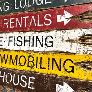 May include: A rustic wooden sign with white painted text on a dark brown background. The sign reads "Hunting Lodge", "Canoe Rentals", "Ice Fishing", "Snowmobiling", and "Outhouse".