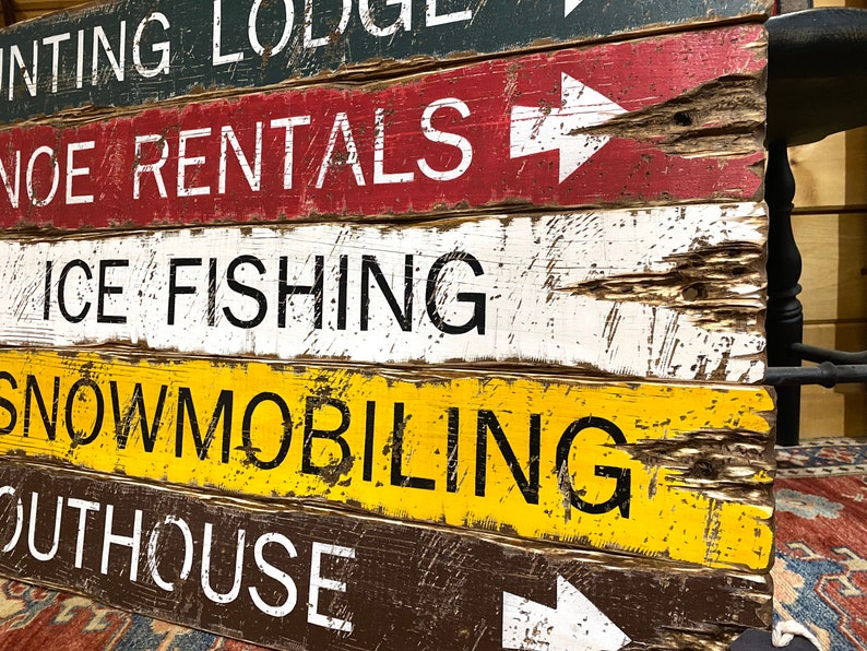 May include: A rustic wooden sign with white painted text on a dark brown background. The sign reads "Hunting Lodge", "Canoe Rentals", "Ice Fishing", "Snowmobiling", and "Outhouse".