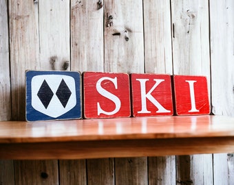 Może przedstawiać: Drewniane klocki z napisem "SKI" z logo ośrodka narciarskiego. Litery są białe na czerwonych klockach, a blok "S" ma niebiesko-białe logo. Klocki są wyświetlane na drewnianej półce na tle drewnianych paneli.