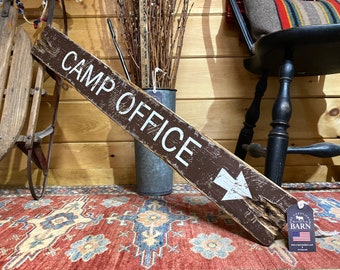 May include: A rustic wooden sign with white lettering that reads "Camp Office" and a white arrow pointing to the right. The sign is hanging on a wooden wall with a red and blue rug in the foreground.