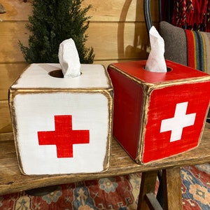 Rustic First Aid wood tissue box cover photographed in a studio setting, ideal for ski cabin décor, lodge bathrooms, beach houses, poolside spaces, and guest rooms. Handcrafted from wood with natural texture, showcasing vintage first aid design and rustic charm for functional home décor