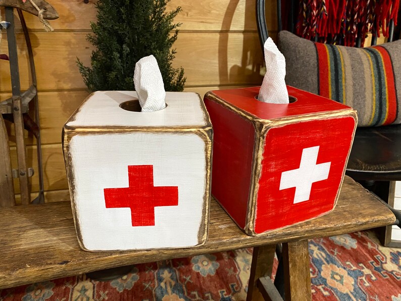 Rustic First Aid wood tissue box cover photographed in a studio setting, ideal for ski cabin décor, lodge bathrooms, beach houses, poolside spaces, and guest rooms. Handcrafted from wood with natural texture, showcasing vintage first aid design and rustic charm for functional home décor