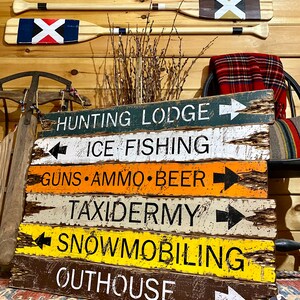 Puede incluir: Carteles de madera con textos como "HUNTING LODGE", "ICE FISHING", "GUNS", "AMMO", "BEER", "TAXIDERMY", "SNOWMOBILING" y "OUTHOUSE". Dos remos con dise&ntilde;os pintados est&aacute;n encima de los carteles. Los carteles son de varios colores.