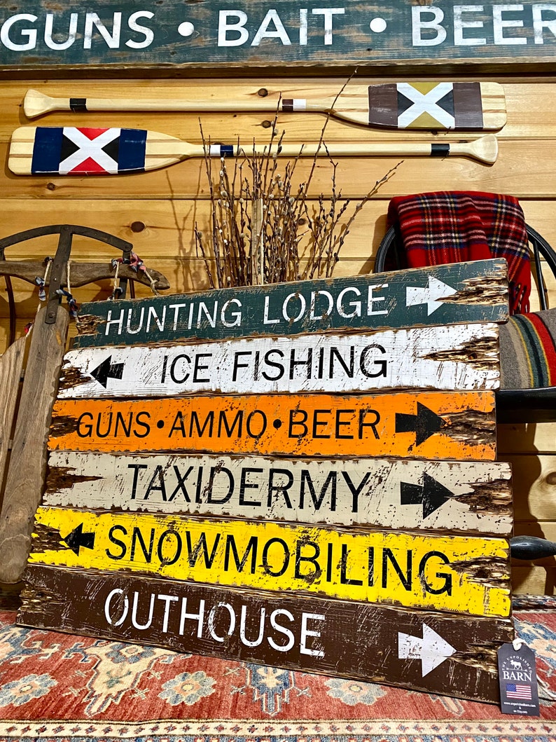 Puede incluir: Carteles de madera con textos como "HUNTING LODGE", "ICE FISHING", "GUNS", "AMMO", "BEER", "TAXIDERMY", "SNOWMOBILING" y "OUTHOUSE". Dos remos con dise&ntilde;os pintados est&aacute;n encima de los carteles. Los carteles son de varios colores.