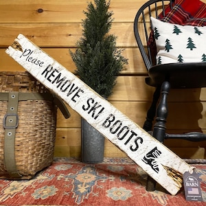 May include: A rustic, white wooden sign with the text "Please REMOVE SKI BOOTS" in black lettering. The sign features a graphic of a ski boot and is propped up on a patterned rug. A small evergreen tree and a woven basket are also visible.