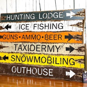 Puede incluir: Un letrero de madera r&uacute;stico con flechas direccionales y texto. El letrero presenta varios destinos como "Hunting Lodge", "Ice Fishing" y "Guns &bull; Ammo &bull; Beer". El letrero tiene un acabado envejecido con una mezcla de colores que incluyen negro, blanco, naranja y amarillo.