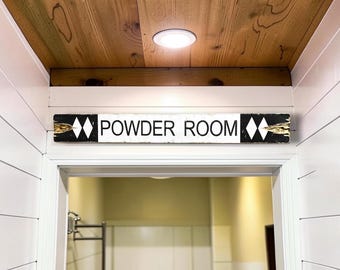 May include: A rectangular wooden sign mounted above a doorway with the text POWDER ROOM in black capital letters on a white background. The ends of the sign are black with white double diamond symbols and gold-toned accents on the edges.