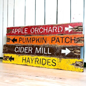 May include: Four wooden signs with arrows pointing to different fall activities. The signs are painted in red, orange, brown, and yellow with white and black lettering. The signs read "Apple Orchard", "Pumpkin Patch", "Cider Mill", and "Hayrides".