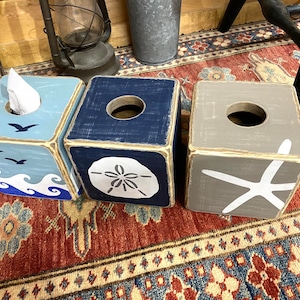 Top view of coastal beach house tissue box covers featuring starfish, sand dollar, and ocean wave designs displayed together, distressed wood finish with relaxed salt-and-sand style, perfect for beach house bathrooms, coastal cottages, lake homes, and vacation rentals, ocean-inspired decor