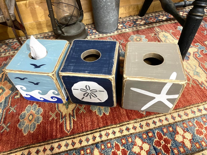 Top view of coastal beach house tissue box covers featuring starfish, sand dollar, and ocean wave designs displayed together, distressed wood finish with relaxed salt-and-sand style, perfect for beach house bathrooms, coastal cottages, lake homes, and vacation rentals, ocean-inspired decor