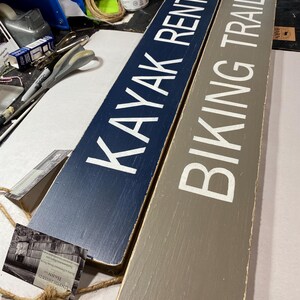 K&ouml;nnte beinhalten: Zwei rechteckige Holzschilder mit wei&szlig;er Beschriftung. Das dunkelblaue Schild zeigt KAYAK RENTALS mit einem Pfeil nach rechts. Das graue Schild zeigt BIKING TRAILS mit einem Pfeil nach rechts. Beide Schilder haben ein Finish und Seil.
