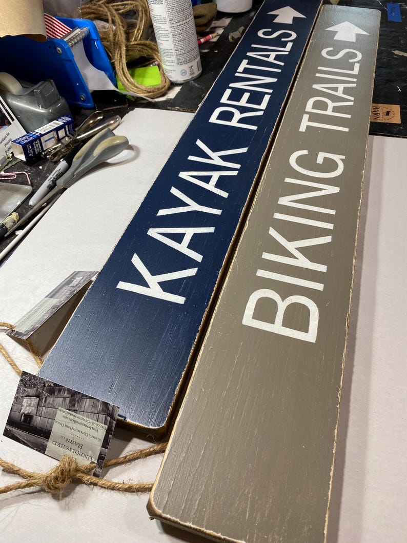K&ouml;nnte beinhalten: Zwei rechteckige Holzschilder mit wei&szlig;er Beschriftung. Das dunkelblaue Schild zeigt KAYAK RENTALS mit einem Pfeil nach rechts. Das graue Schild zeigt BIKING TRAILS mit einem Pfeil nach rechts. Beide Schilder haben ein Finish und Seil.