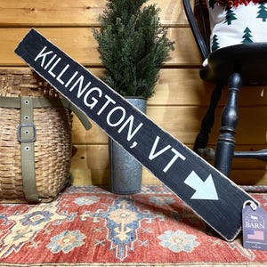 May include: Black wooden sign with white text that reads "Killington, VT" and a white arrow pointing to the right. The sign is on a red and cream patterned rug. A small tag with the text "The American Barn" is attached to the sign.