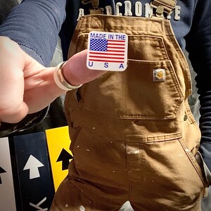 May include: A person wearing brown overalls with a pocket on the right side. The overalls have a white sticker on the left side that says "Made in the USA" with an American flag graphic.