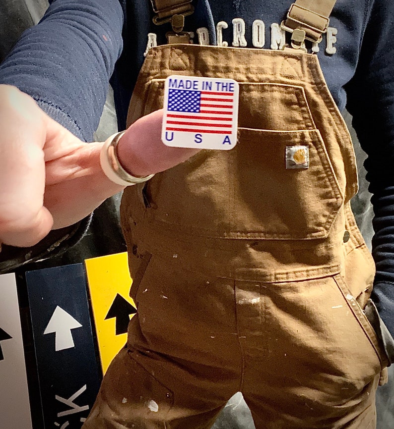 May include: A person wearing brown overalls with a pocket on the right side. The overalls have a white sticker on the left side that says "Made in the USA" with an American flag graphic.
