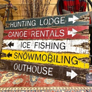 May include: A rustic wooden sign with five rows of text and arrows pointing in different directions. The text reads: "Hunting Lodge", "Canoe Rentals", "Ice Fishing", "Snowmobiling", and "Outhouse".