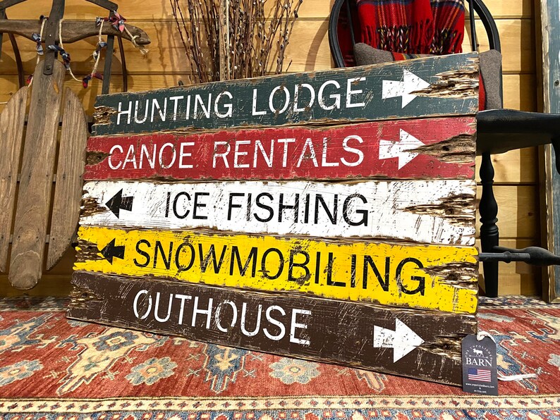 May include: A rustic wooden sign with five rows of text and arrows pointing in different directions. The text reads: "Hunting Lodge", "Canoe Rentals", "Ice Fishing", "Snowmobiling", and "Outhouse".