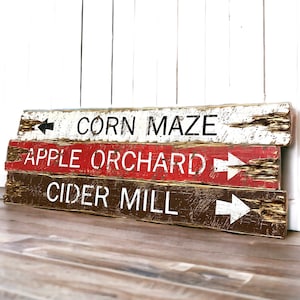 May include: Three wooden signs with white painted arrows and black text. The top sign reads "CORN MAZE", the middle sign reads "APPLE ORCHARD", and the bottom sign reads "CIDER MILL".