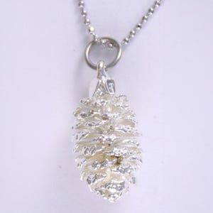 May include: A silver-colored necklace featuring a detailed pinecone pendant. The pendant is intricately designed with textured scales, suspended from a delicate chain. The necklace is a nature-inspired piece, suitable for everyday wear.