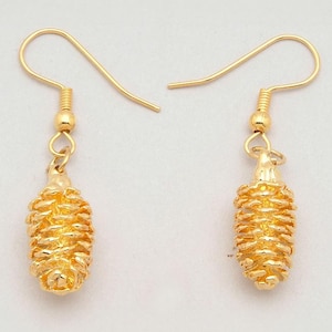 Puede incluir: Un par de pendientes dorados. Cada pendiente presenta un dije de piña texturizado y detallado, suspendido de un gancho simple. Los pendientes están diseñados con un acabado metálico cálido y se muestran sobre un fondo blanco.