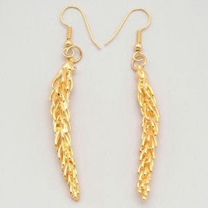 May include: A pair of gold-colored earrings. Each earring features a long, textured, wheat-like design suspended from a simple hook. The earrings are displayed against a plain background, highlighting their intricate details and warm metallic hue.