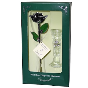 May include: A single black rose dipped in platinum, preserved in a clear glass vase. The rose and vase are displayed in a green box with the text "Real Rose Dipped in Platinum" and "Living Gold!"