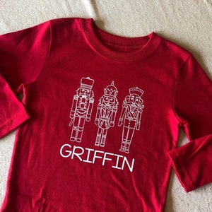 May include: Red long-sleeve shirt with a white design of three nutcracker figures and the name "GRIFFIN" printed below.
