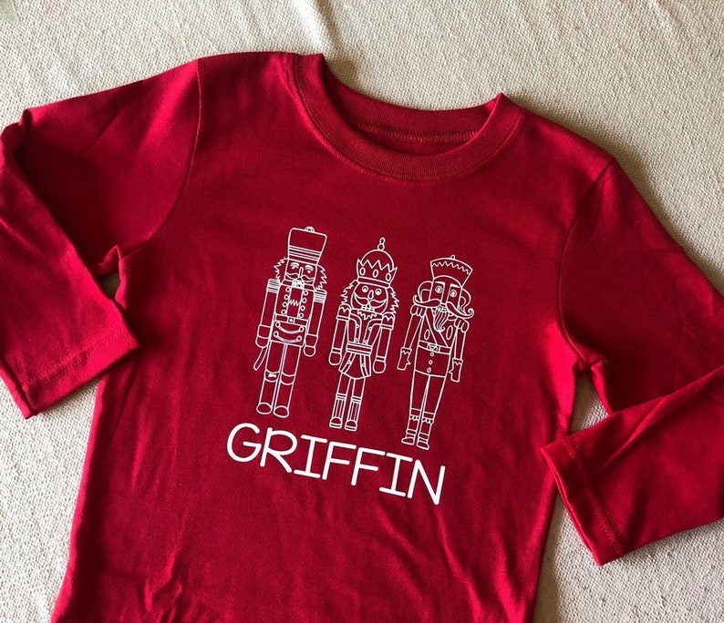 May include: Red long-sleeve shirt with a white design of three nutcracker figures and the name "GRIFFIN" printed below.
