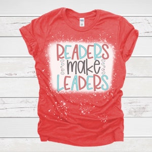 May include: A red t-shirt with the words "READERS make LEADERS" in a colorful design. The text is in a white rectangle with a distressed, speckled effect. The shirt has rolled-up sleeves.