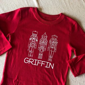 May include: Red long-sleeve shirt with a white design of three nutcracker figures and the name "GRIFFIN" printed below.
