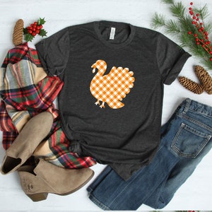 May include: A dark gray t-shirt with a white and orange gingham turkey graphic. The shirt is laid out on a white surface with a plaid scarf, brown boots, and blue jeans.