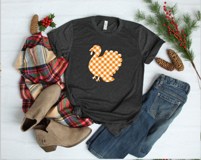 May include: A dark gray t-shirt with a white and orange gingham turkey graphic. The shirt is laid out on a white surface with a plaid scarf, brown boots, and blue jeans.